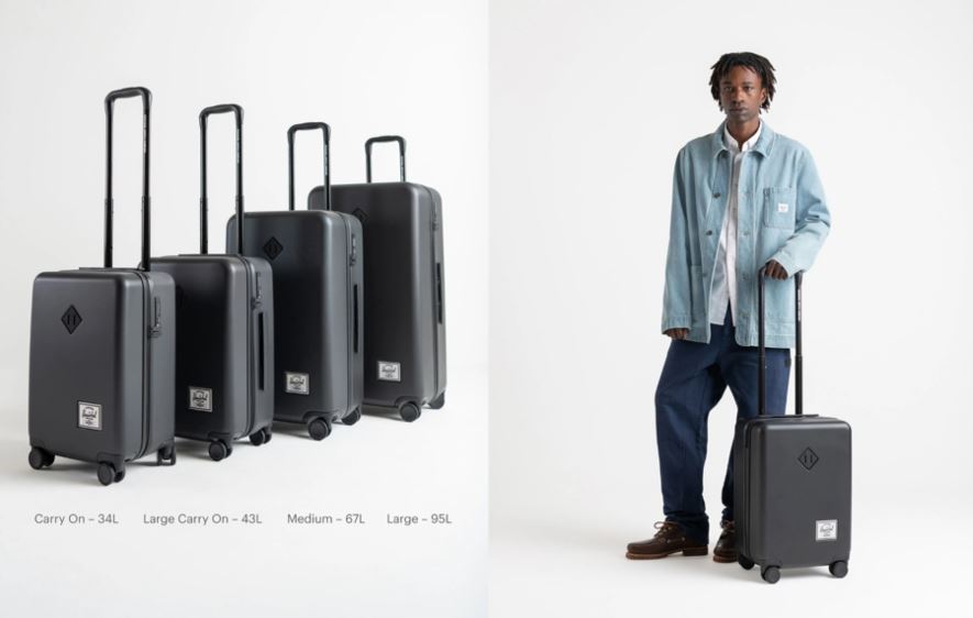 Man in a light blue jacket and white shirt and dark blue jeans standing next to a small piece of luggage on wheels and also there is another group of luggae, five pieces of them and they are all different a sizes and all have wheels.